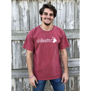 Young male wearing a firecracker red short sleeve tee with the Kayak design by Chillwater. The back resembles a kayaker floating down a river covered by trees.
