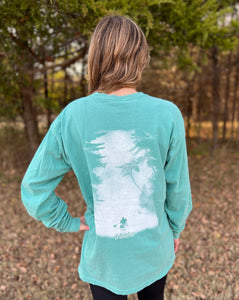 Young female wearing a tropical timid teal long sleeve tee with the Kayak design by Chillwater.The back resembles a kayaker floating down a river covered by trees.