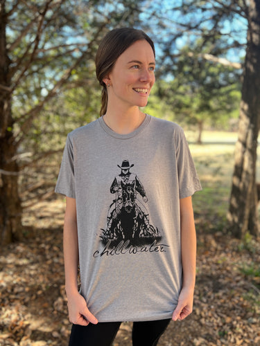 Young female wearing a barn metal grey short sleeve tee in the Rawhide design by Chillwater. The front pictures a cowboy riding horseback.