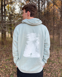 Young male wearing a soft cypress green sweatshirt hoodie with the Kayak design by Chillwater. The back resembles a kayaker floating down a river covered by trees.
