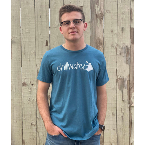 Young male wearing a stone blue short sleeve tee with the Kayak design by Chillwater. The back resembles a kayaker floating down a river covered by trees.