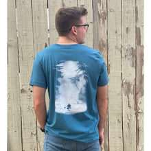 Young male wearing a stone blue short sleeve tee with the Kayak design by Chillwater. The back resembles a kayaker floating down a river covered by trees.
