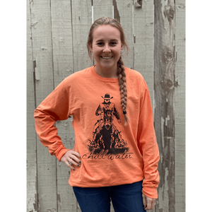 Young female wearing a sunrise orange long sleeve tee in the Rawhide design by Chillwater. The front pictures a cowboy riding horseback.