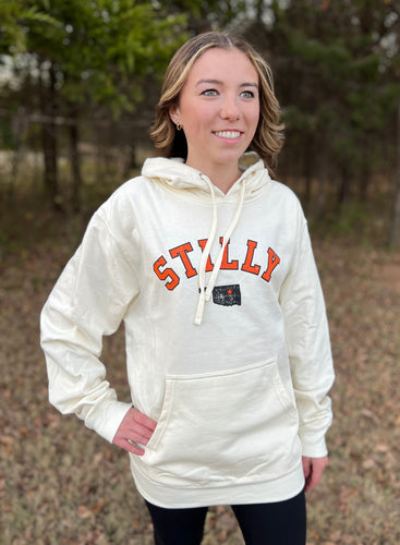 Young female wearing a washed ivory hooded sweatshirt in the Stilly design by Chillwater Apparel. The front shows the words “Stilly” with the state of Oklahoma and a star over Stillwater.