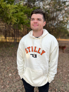 Young male wearing a washed ivory hooded sweatshirt in the Stilly design by Chillwater Apparel. The front shows the words “Stilly” with the state of Oklahoma and a star over Stillwater.