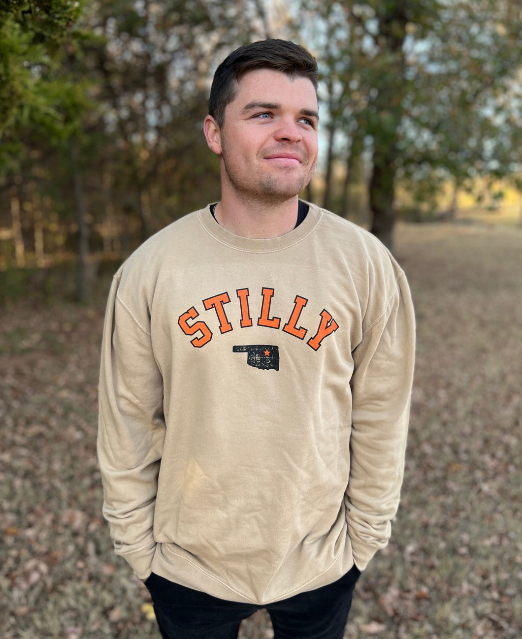 Young male wearing a cobblestone khaki sweatshirt in the Stilly design by Chillwater Apparel. The front shows the words “Stilly” with the state of Oklahoma and a star over Stillwater.