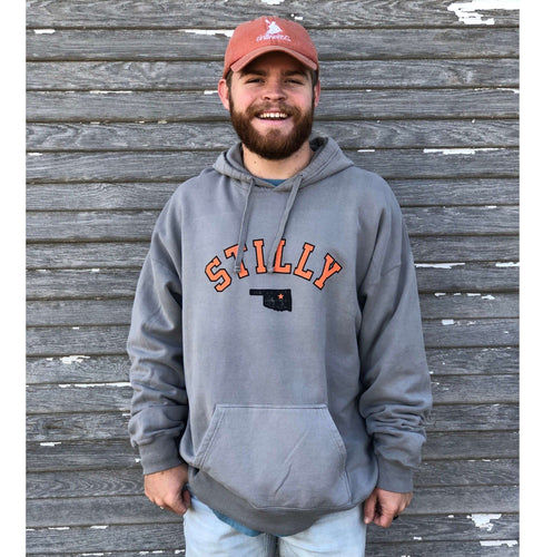 Grey comfort stretch long sleeve hoodie sweatshirt. On the center of the hoodie is the chiillwater stilly logo in orange lettering with black outline and small black Oklahoma with orange star over stillwater location.