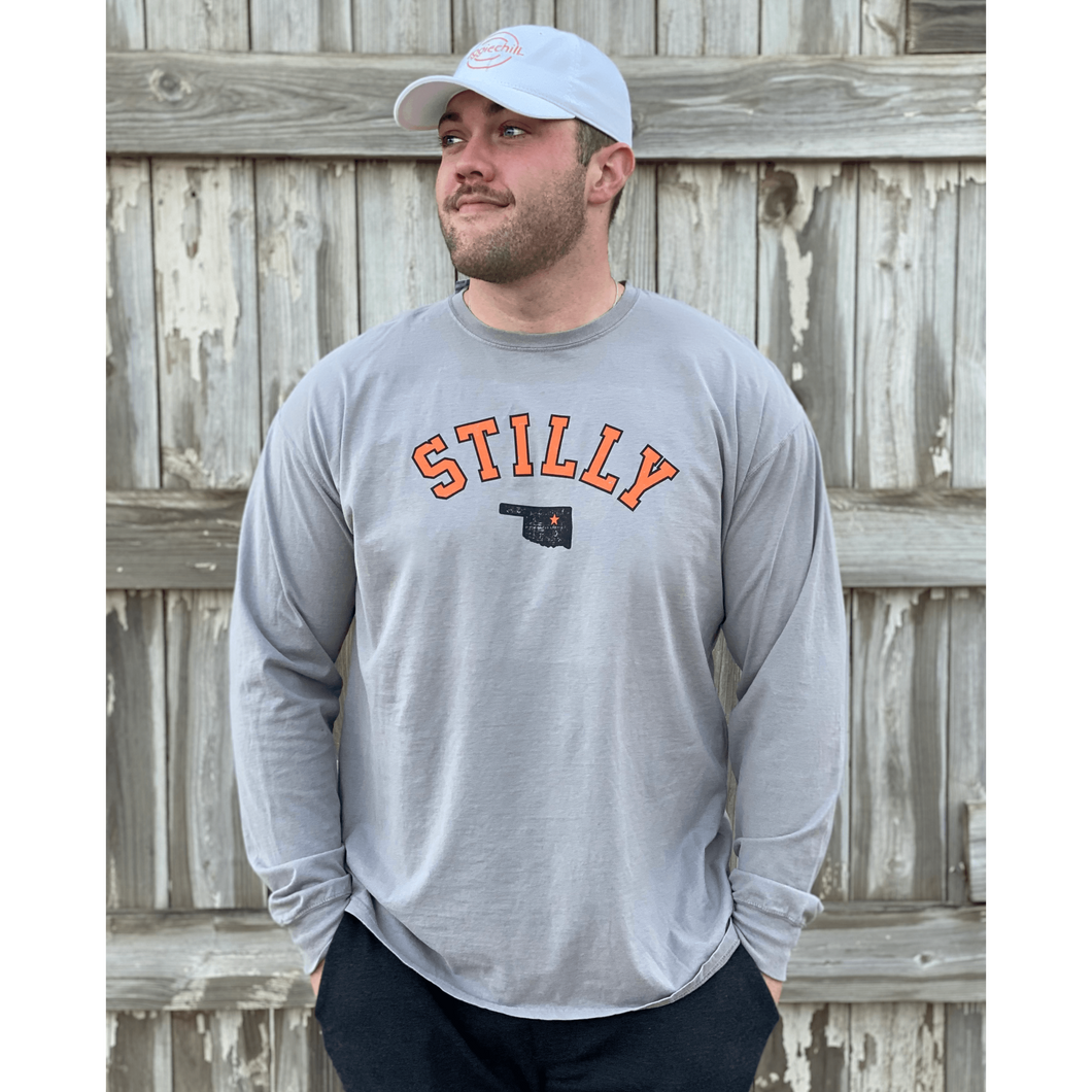 Young male wearing a lava grey long sleeve tee in the Stilly design by Chillwater Apparel. The front shows the words “Stilly” with the state of Oklahoma and a star over Stillwater.